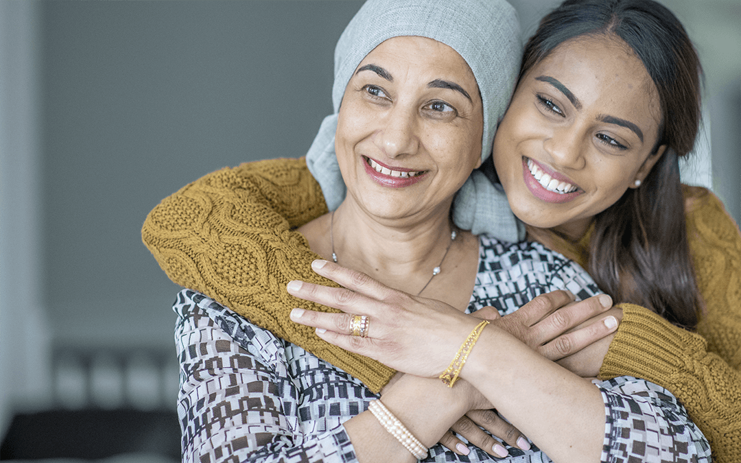 Photo of a senior woman is dressed with a head scarf on while her daughter hugs her from behind. Photo d'une femme âgée vêtue d'un foulard pendant que sa fille la serre dans ses bras par derrière.